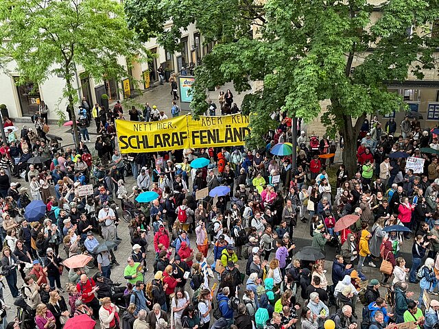 Eine große Menschenmenge protestiert im Freien mit Schildern und einem gelben Transparent unter Bäumen und Sonnenschirmen.
