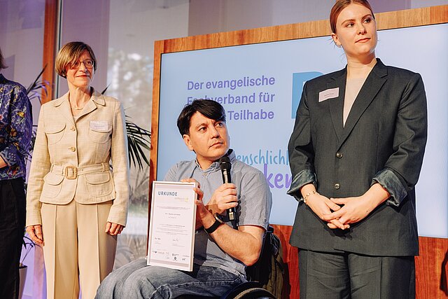 Ein Mann im Rollstuhl hält eine Urkunde und ein Mikrofon in der Hand, während zwei Frauen neben ihm bei einer Veranstaltung stehen.