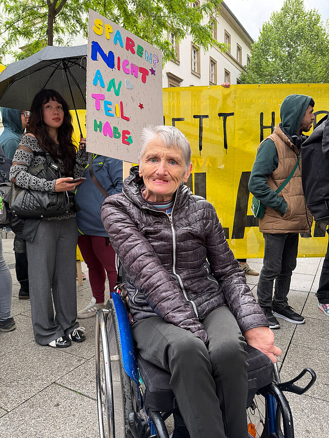 Ältere Frau im Rollstuhl bei einer Demonstration, mit Menschen und einem bunten Schild hinter einem gelben Banner.