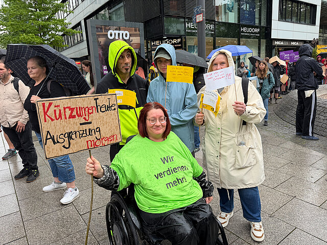 Vier Menschen halten bei einer verregneten Demonstration Schilder mit der Forderung nach höheren Löhnen; einer sitzt im Rollstuhl.