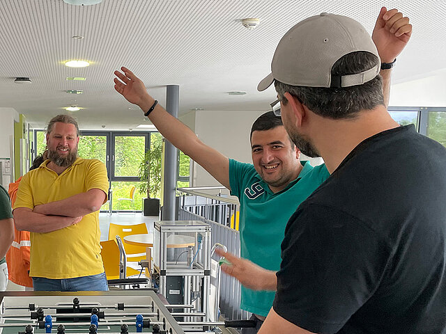 Zwei Männer feiern beim Kickerspiel, einer mit siegreich erhobenen Armen, die anderen schauen zu und lächeln in einem hellen Raum.