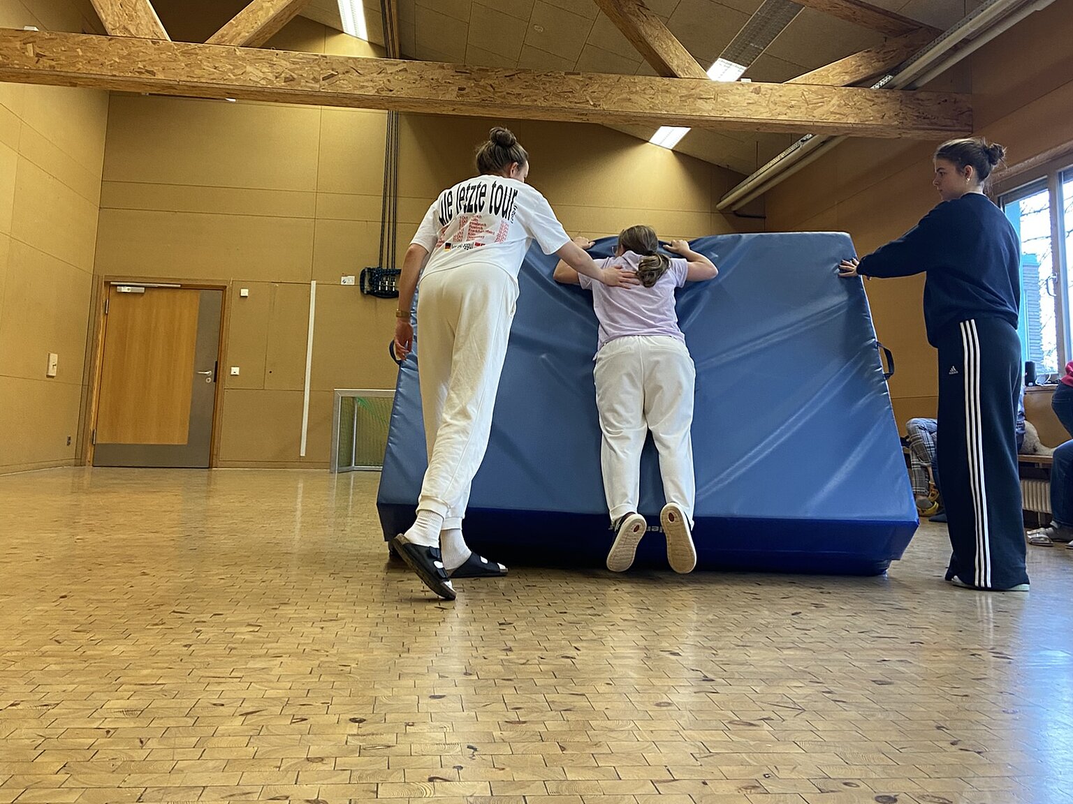Drei Personen bewegen in einer Turnhalle eine große blaue Matte; zwei schieben, einer führt von der Seite.