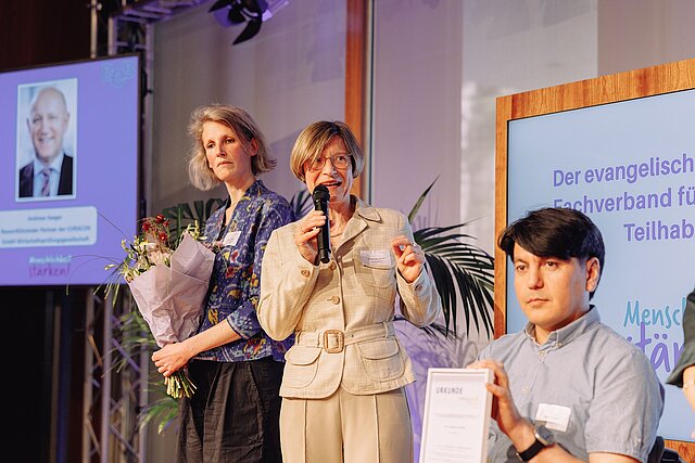 Eine Frau spricht in ein Mikrofon, während eine andere Blumen hält; ein Mann sitzt mit einer Urkunde in der Hand.