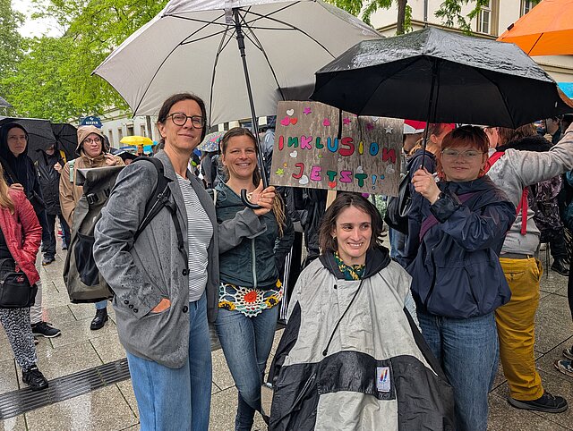Vier Personen halten bei einer regnerischen Demonstration im Freien Regenschirme und ein buntes Schild mit der Aufschrift "Inklusion Jetzt!".